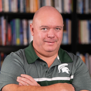 Headshot of Phil Rumrill wearing a green Spartan polo, standing in front of a bookshelf