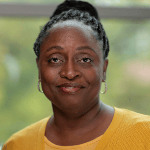 Angela Smith wearing a yellow sweater. There is greenery in the background. 