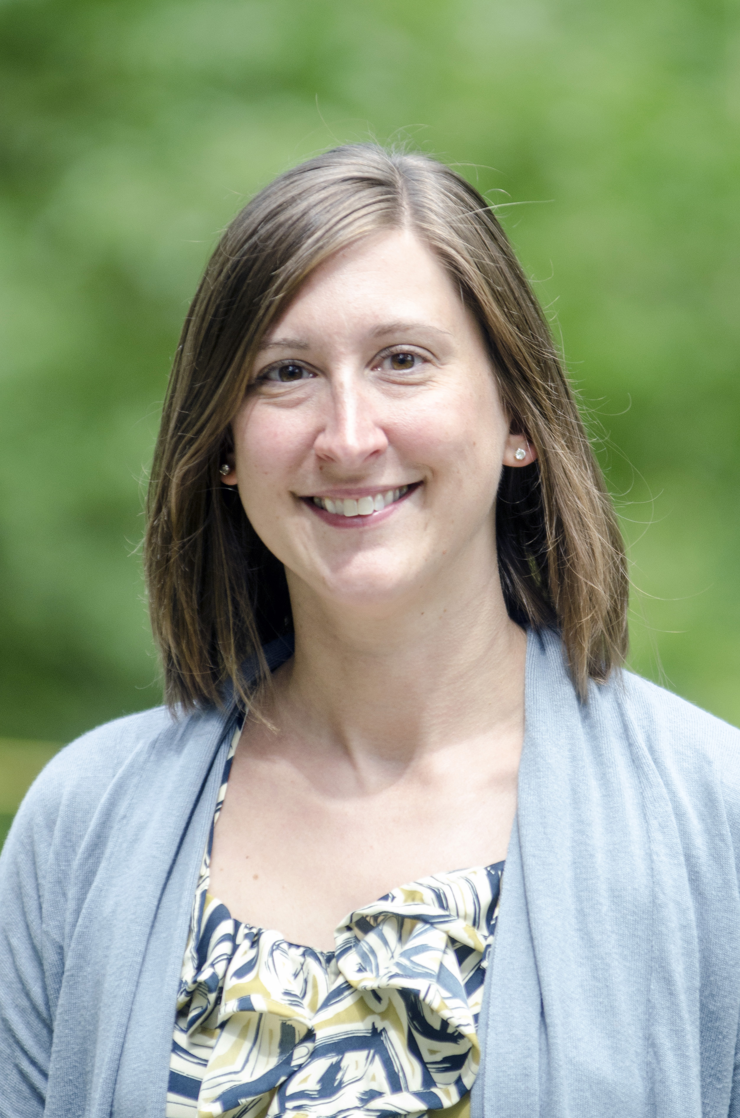 Jennifer VanDerHeide wearing a light blue sweater and blue/green patterned shirt. There is greenery in the background. 