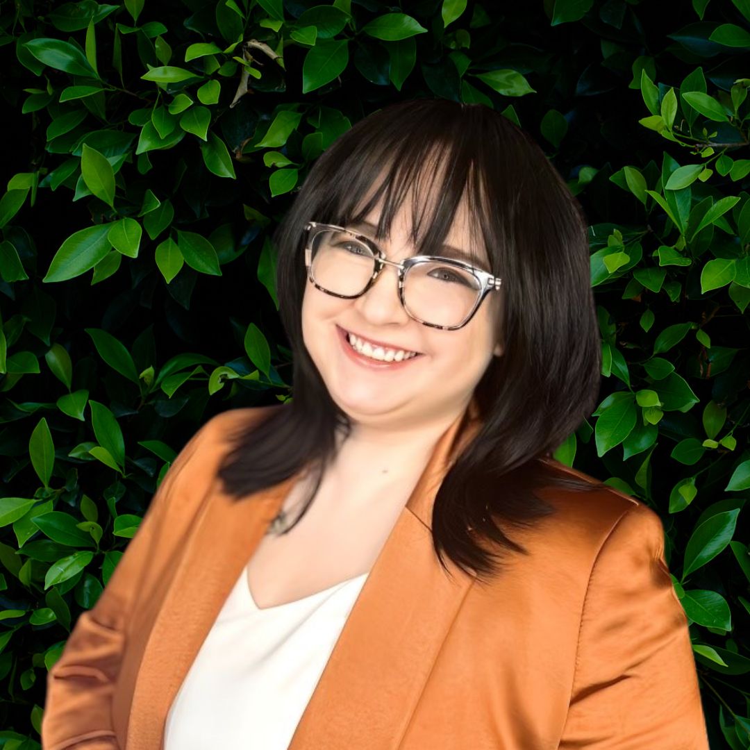 Voort Uher smiles for the camera while standing in front of a bush lush with dark green leaves. Voort Uher wears a burnt orange blazer over a cream colored top and dark-rimmed glasses. Voort Uher's hair is dark; Voort Uher has shoulder-length hair and bangs.