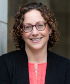 Tanya Wright headshot. She wears a pink shirt under a dark blazer. Her curly brown hair is shoulder length; she wears dark-rimmed glasses.