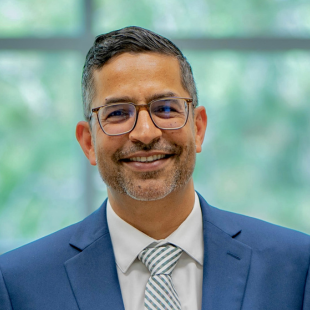 Aman Yadav smiles for the camera while standing outside in front of a bush lush with green leaves. Yadav wears a blue, pink and green plaid shirt under a royal blue blazer. Yadav wears dark-rimmed glasses. Yadav's black hair is short.
