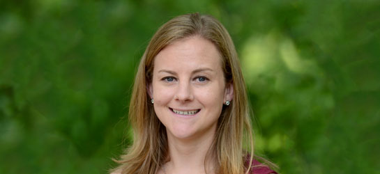Marisa Fisher smiles for the camera while standing outdoors in front of bushes lush with green leaves. Fisher wears a red sweater. Fisher's blonde hair is long and falls past the shoulders.