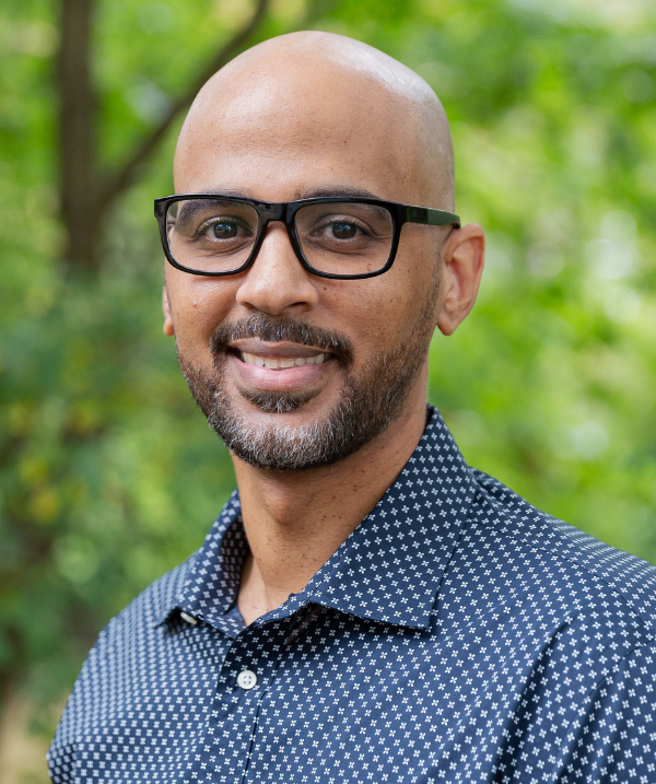 Terry Flennaugh smiles for the camera while standing outdoors in front of a bush lush with green leaves. Terry wears a navy blue button down with white polka dots and dark-rimmed glasses.