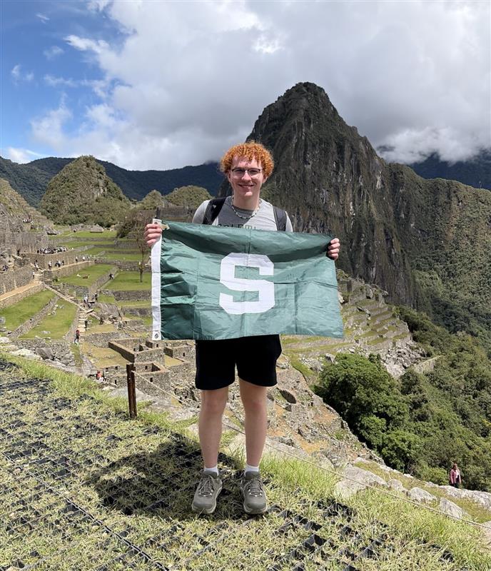 Jagger Wraalstad stands on top of a mountain at Machu Picchu and holds a green and white MSU flag. 