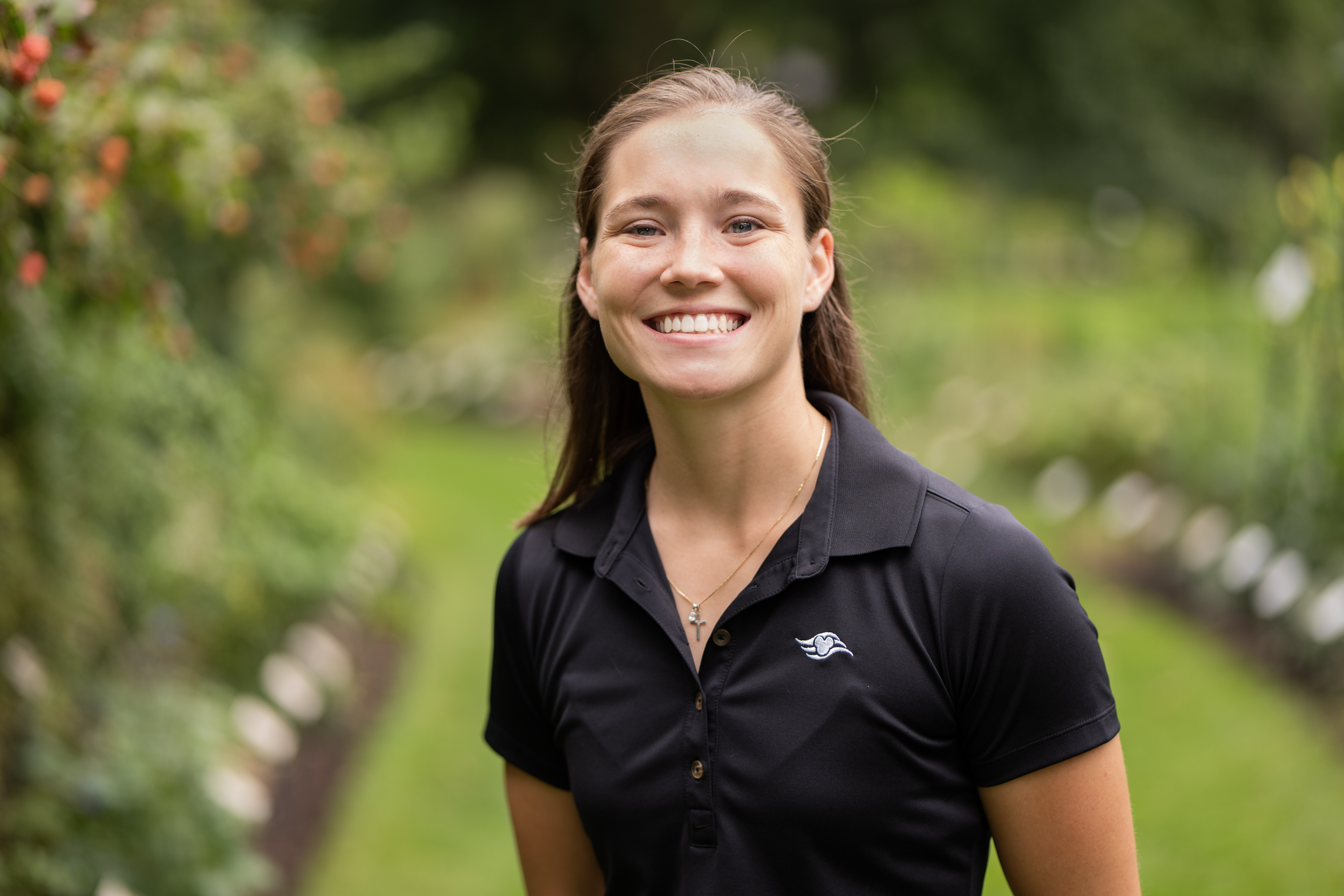Corinne Zimmerman Hobbs smiles in an outdoor setting. Corinne wears a black polo tee-shirt with a small white Disney logo in the torso. Corinne has longe brown hair, pulled half back.