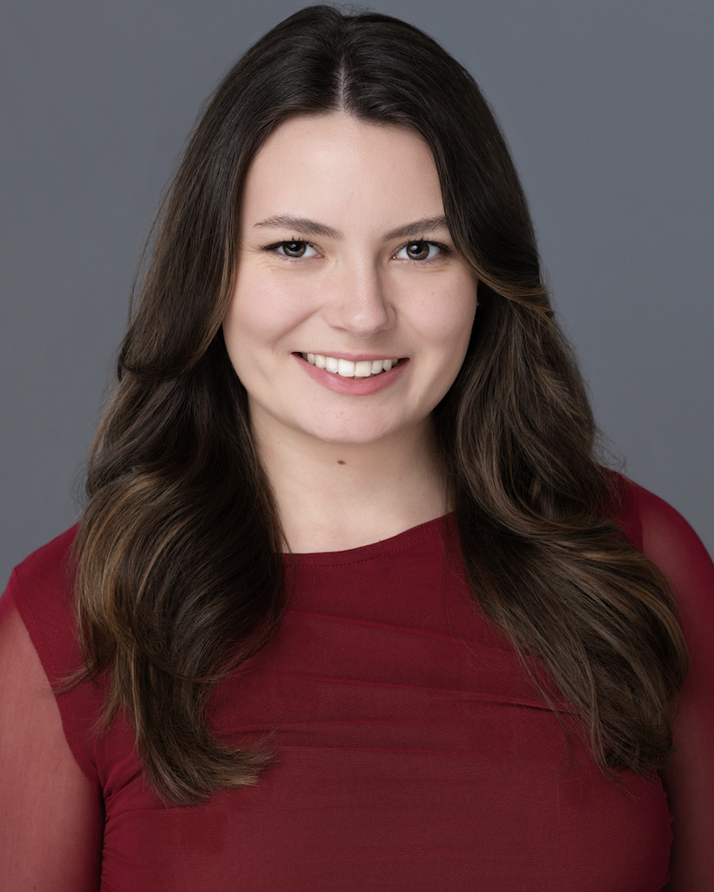 Lilian Klein smiles for the camera against a neutral gray backdrop. Klein wears a dark red, long-sleeved blouse. Klein has long brown hair that is curled at the end.
