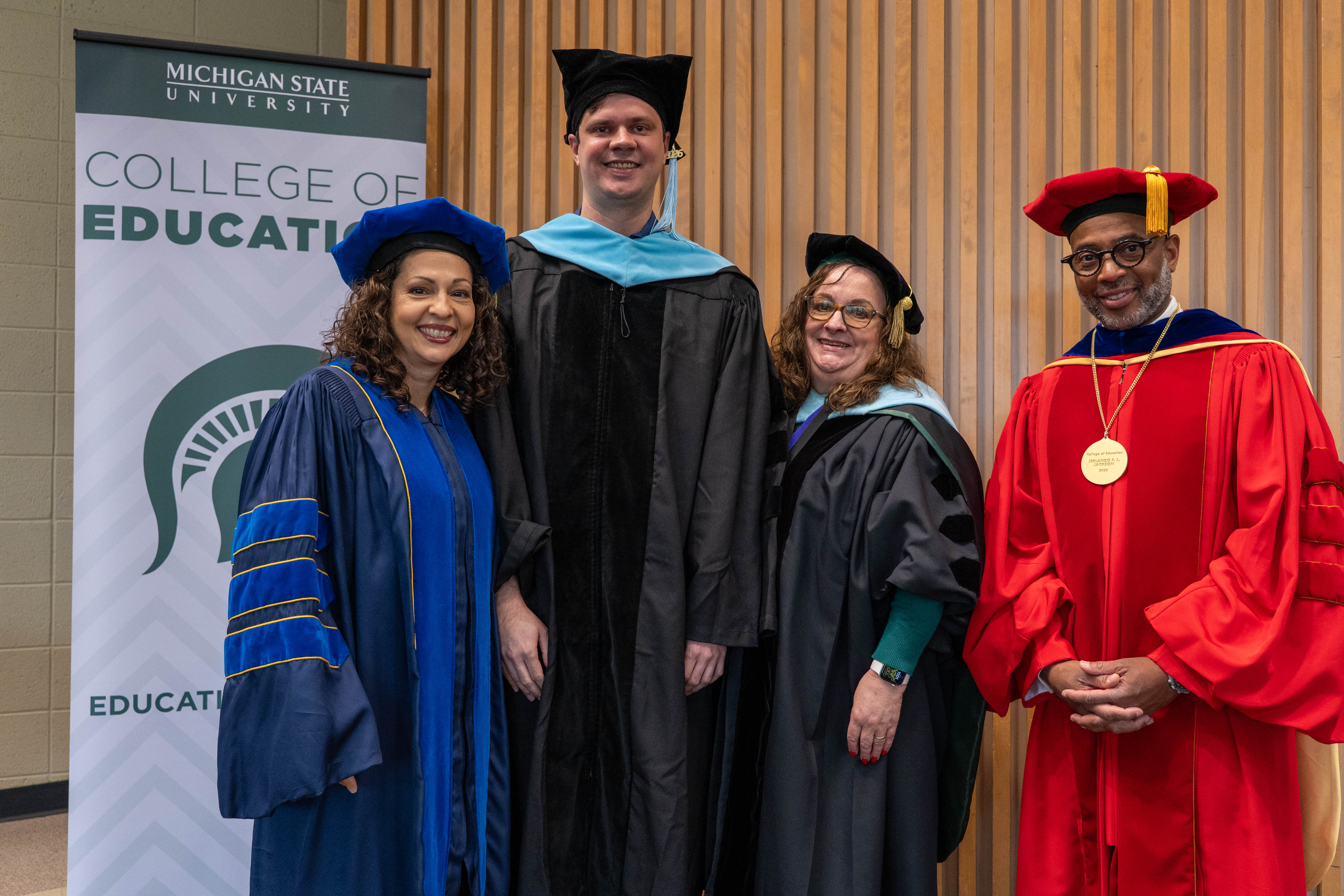 Four Spartans stand side-by-side in various colored graduation regalia. All are in the Erickson Hall Kiva for a doctoral convocation. A sign for the College of Education is behind the Spartans.