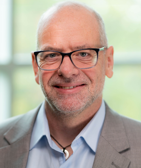 Matt Koehler smiles for the camera while standing in front of a set of windows. Koehler wears a light blue button-down shirt under a light gray blazer as well as wire-rimmed glasses