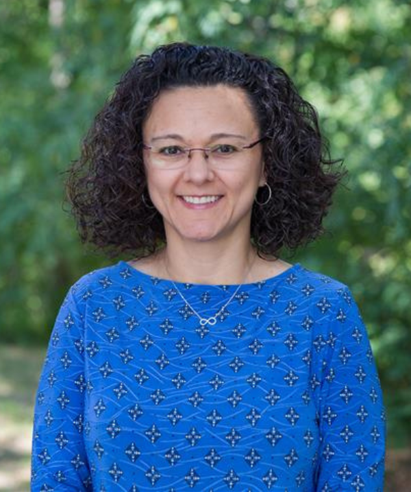 Patricia Marin smiles for the camera while standing outdoors in front of a bush lush with green leaves. Marin wears a blue top with black and white patterns on it, a silver infinity necklace, silver hoop earrings and wire-rimmed glasses. Marin's black hair is curly and about shoulder-length.