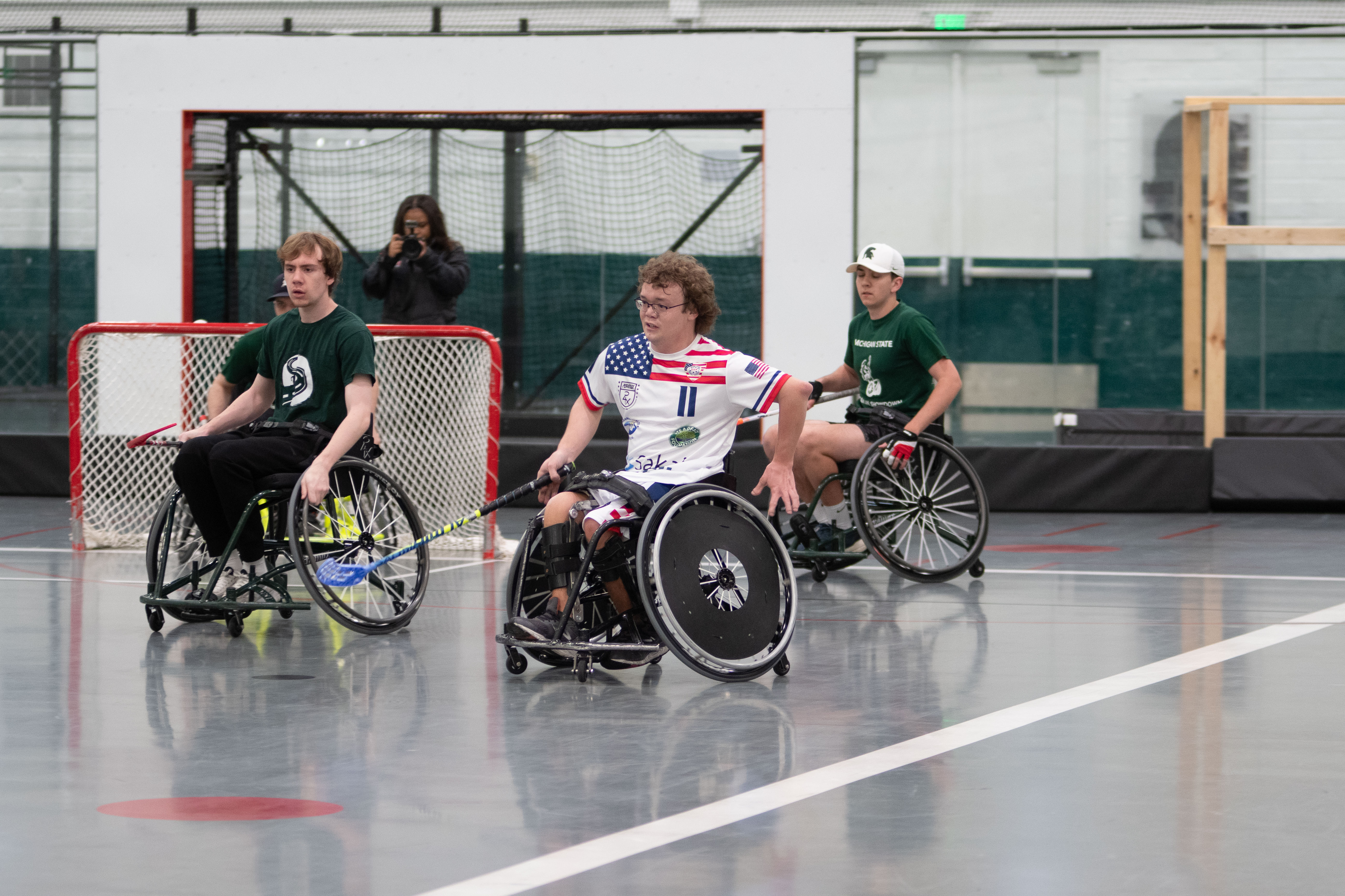 MSU Floorball team competes during a game. MSU athlete, wearing a red white and blue jersey, races toward a ball while holding a stick with a blue handle. Players from the opposing team wear dark-green shirts and surround the MSU player.