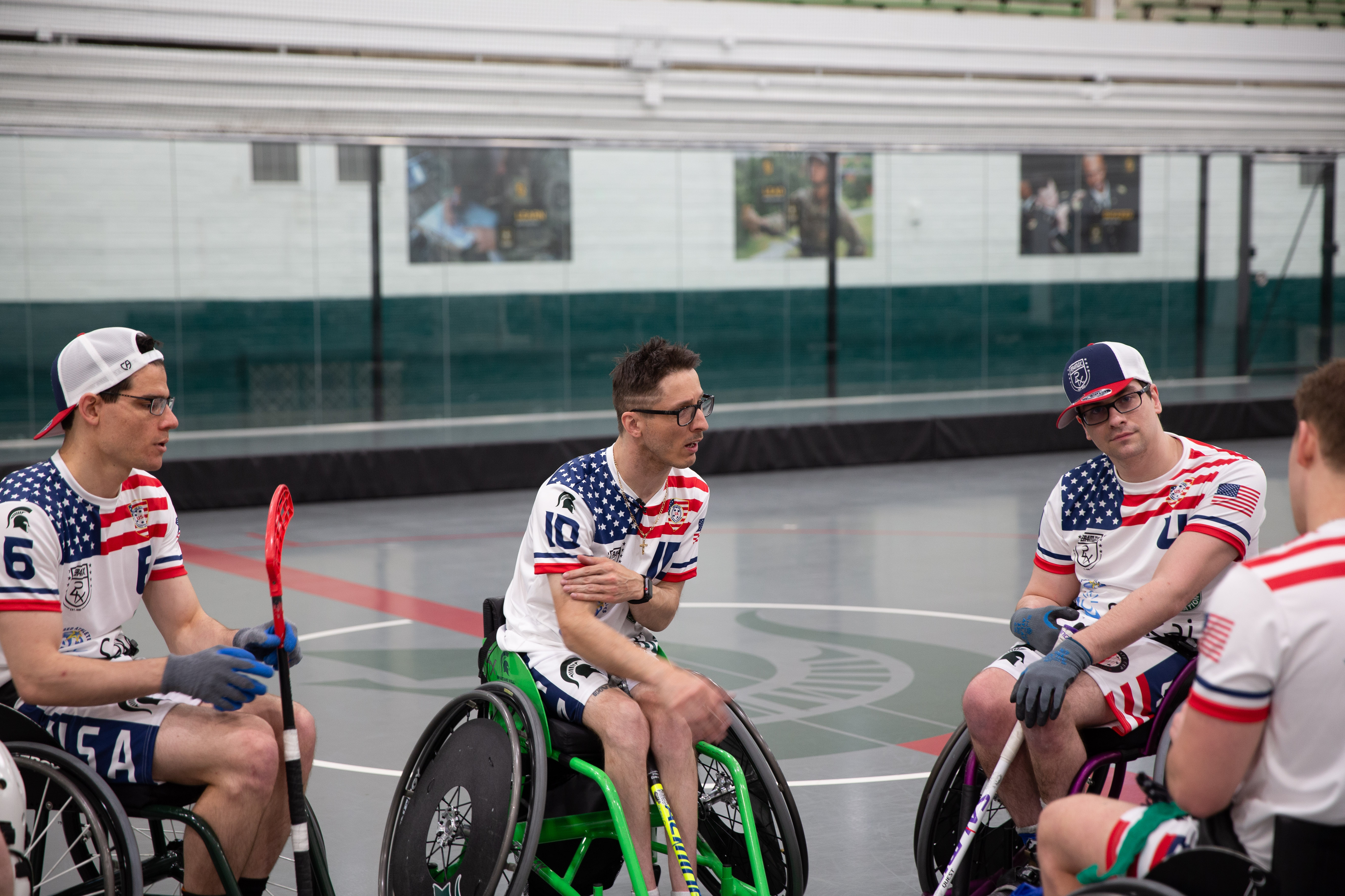 Piotr Pasik and colleagues are on the adaptive floorball court, huddled in conversation. The four individuals wear red, white and blue jerseys. Most wear gloves on their hands. All are seated in adaptive wheelchairs used for rugby, floorball and other adaptive sports.