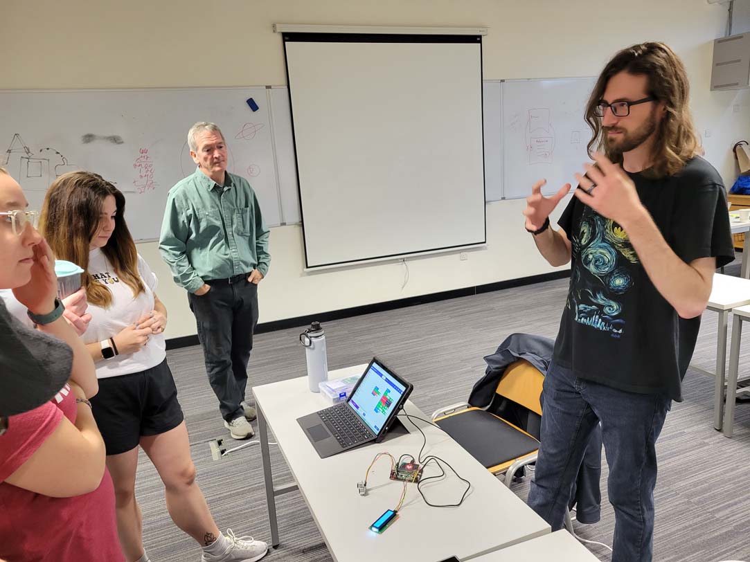 Spartan educator demonstrates a learning topic while standing nearby an open and active laptop. Students listen in.