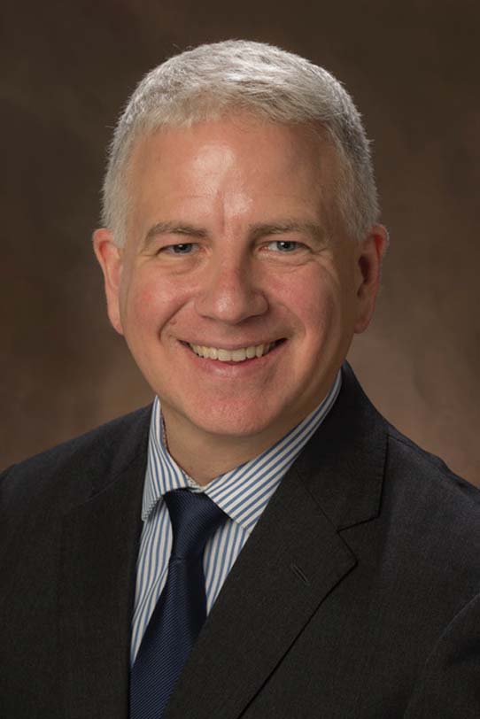 Jeffrey Wanko poses for a photo against a neutral brown background. Wanko wears a dark suit. Wanko's hair is gray and cropped close to the head.