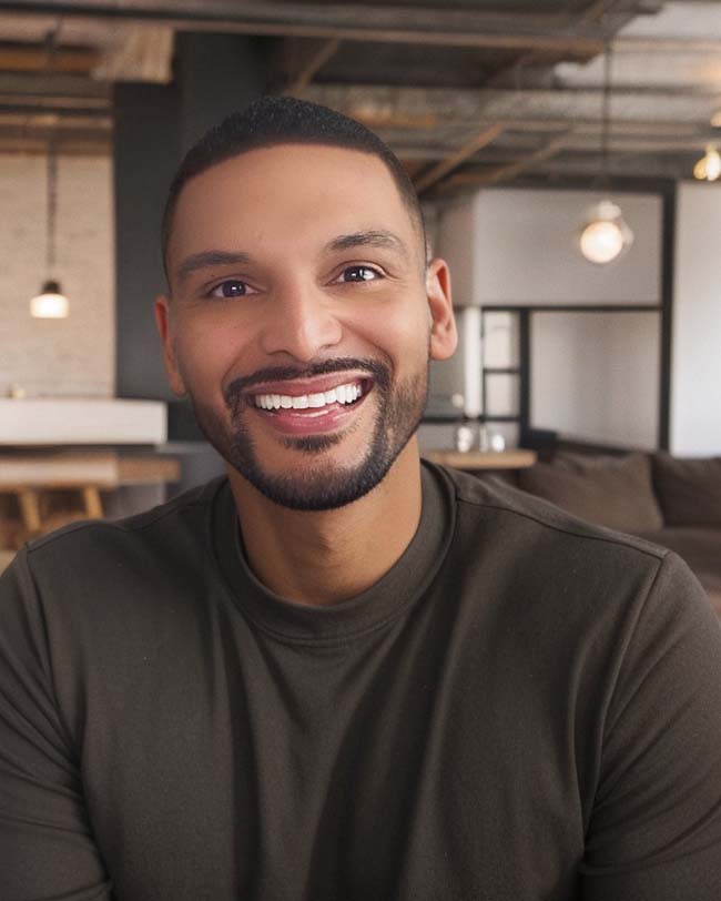 Stephen Santa-Ramirez poses for the camera inside of an office-like setting. Santa-Ramirez wears a dark shirt. Santa-Ramirez has a dark beard and dark hair.