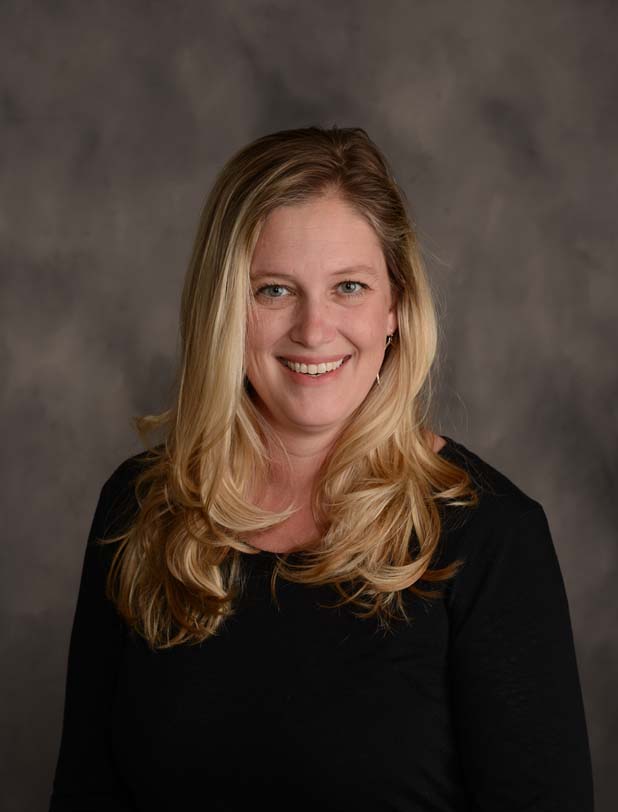 Tami Mannes poses in front of a neautral brown backgorund. Tami wears a dark blouse. Tami's blonde hair is curled and falls past the shoulders.