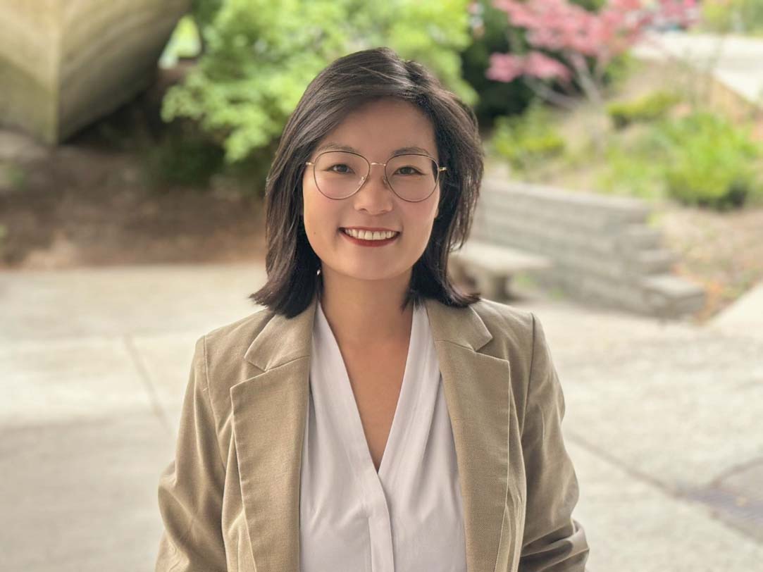 Tingting Li poses for a photo in an outside venue on a sidewalk. TingTing wears a white blouse under a beige jacket. TingTing wears glasses. TingTing's black hair is shoulder-length.