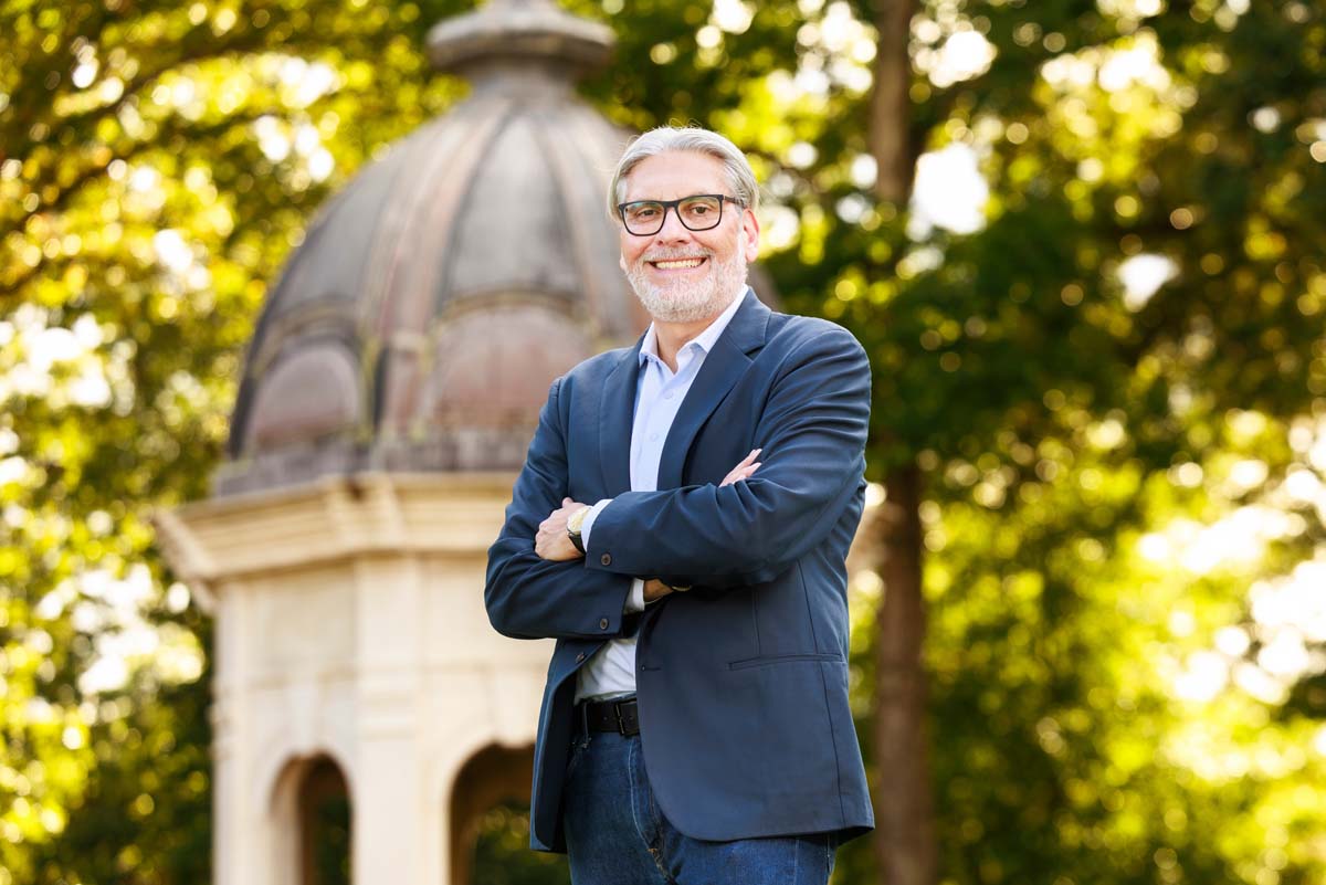 Matthew Militello poses for a photo at an outside venue across from a stone archway. Miliello wears a dark suit. Militello has glasses. Militello has gray hair and a gray beard. Photo by Steven Mantilla.