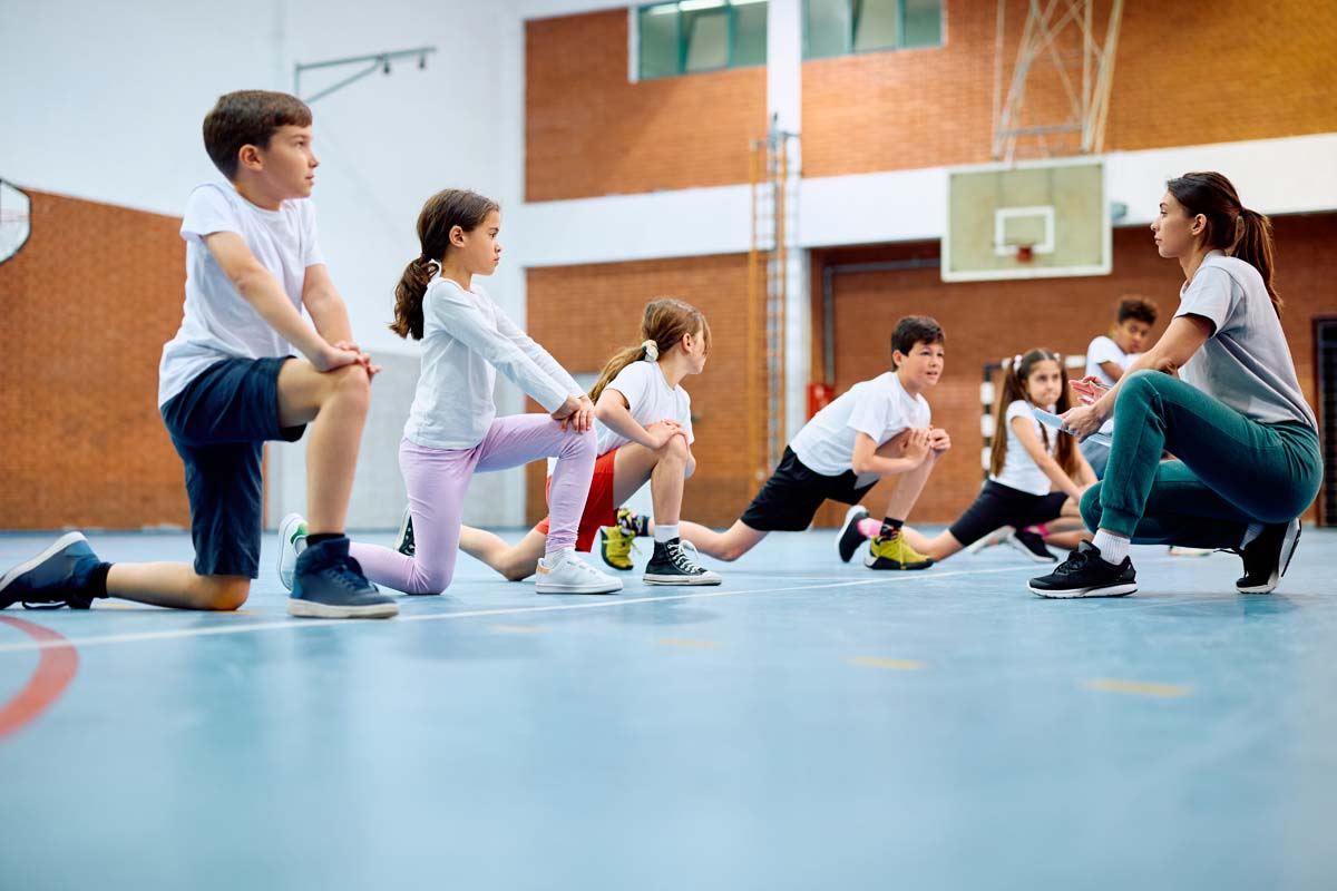 Group of children exercise during a physical education class. A teacher kneels nearby.