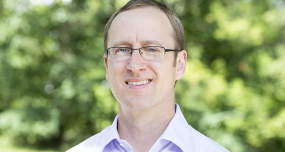 Nick Myers headshot. Myers stands outside against green foliage and smiles toward the camera. Myers wears a light-colored button down. Myers has wire-rimmed glasses and blonde hair cropped close to the head.
