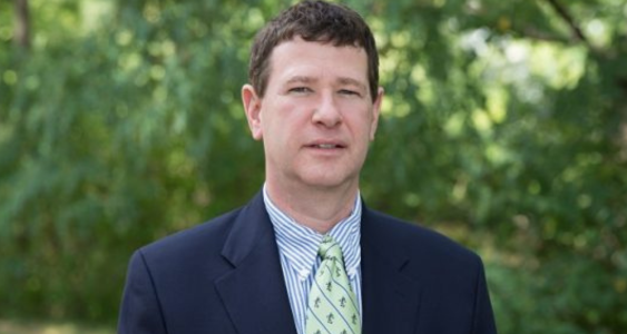 Matthew Wawrzynski headshot. Wawrzynski stands against a backdrop of trees and smiles toward the camera. Wawrzynski wears a dark blue blazer over a light blue button-down and green patterned tie.