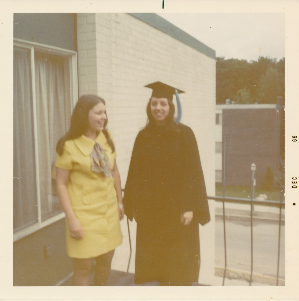 Gwen stands on an outdoor balcony in black commencement regalia with a light blue tassel on cap. Gwen stands near another, dressed in yellow. Along the side of the image, 