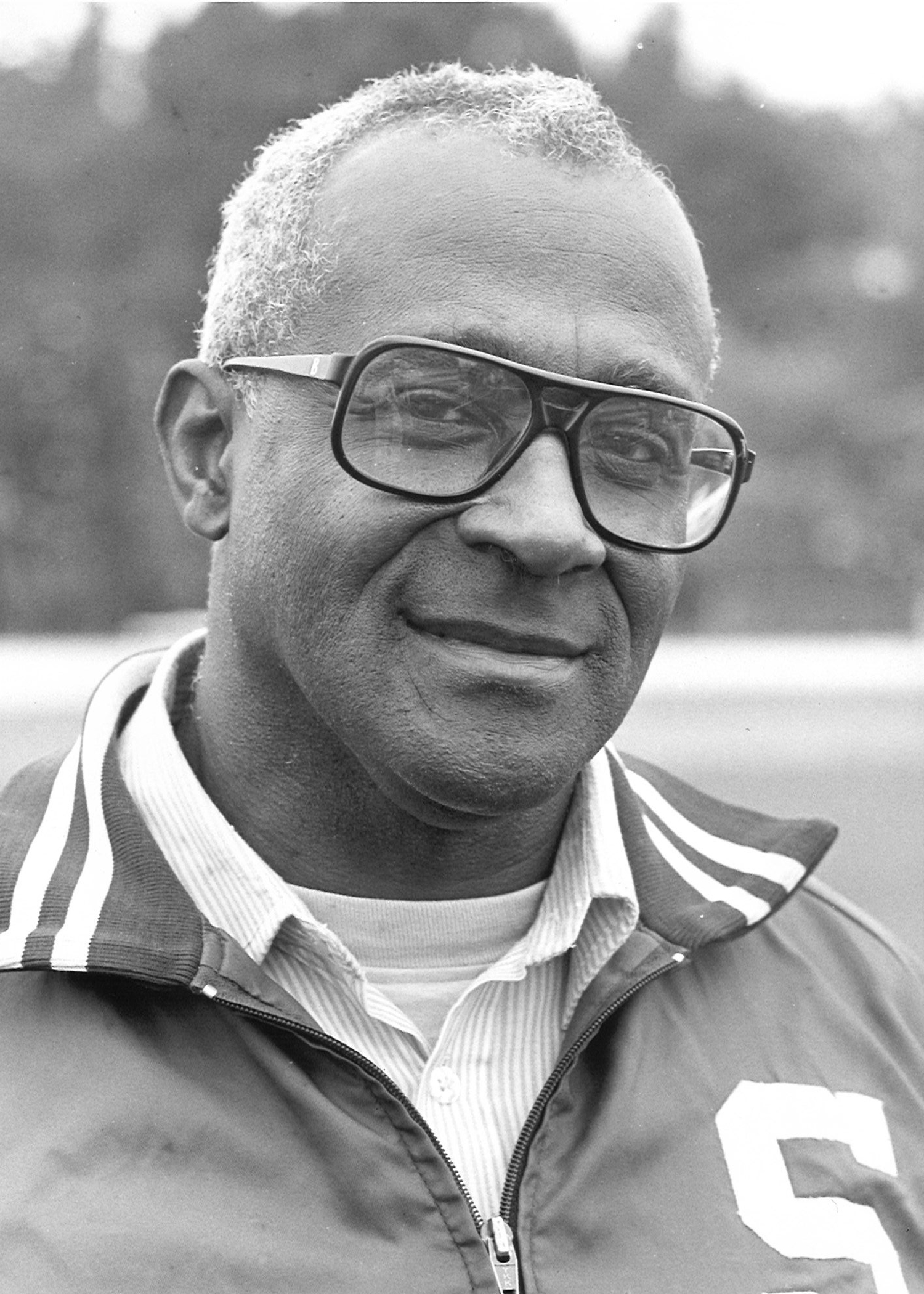 Jim Bibbs smiles toward the camera while standing in an outdoor setting in a black and white image. The image is focused on Bibbs' torso; Bibbs wears an MSU jacket, with most of a Spartan 