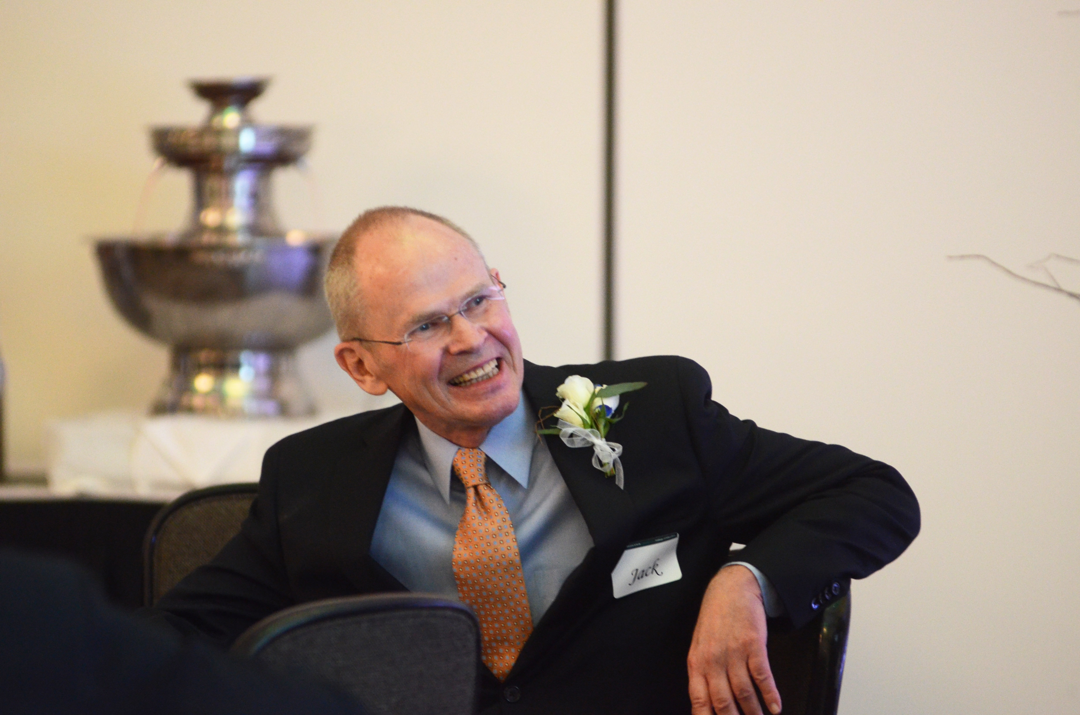 Jack Schwille turns in his chair and smiles off-camera. Jack is wearing a blue button-down shirt and orange tie with a black blazer. Jack wears a white rose boutonnière and a nametag with