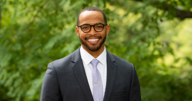 Terrance Burgess smiles for the camera in an outdoor setting in front of a bush lush with green leaves. Burgess wears a lavender tie over a white button-down shirt, as well as a black blazer and dark-rimmed glasses. Burgess's black hair and beard are short.