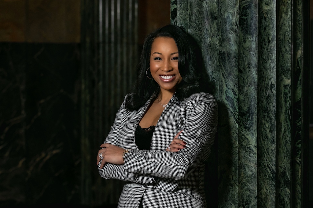 LaToya Thompson leans against a green marble ionic column. Thompson wears a blazer with a black and white checked pattern. Thompson has shoulder-length black hair.