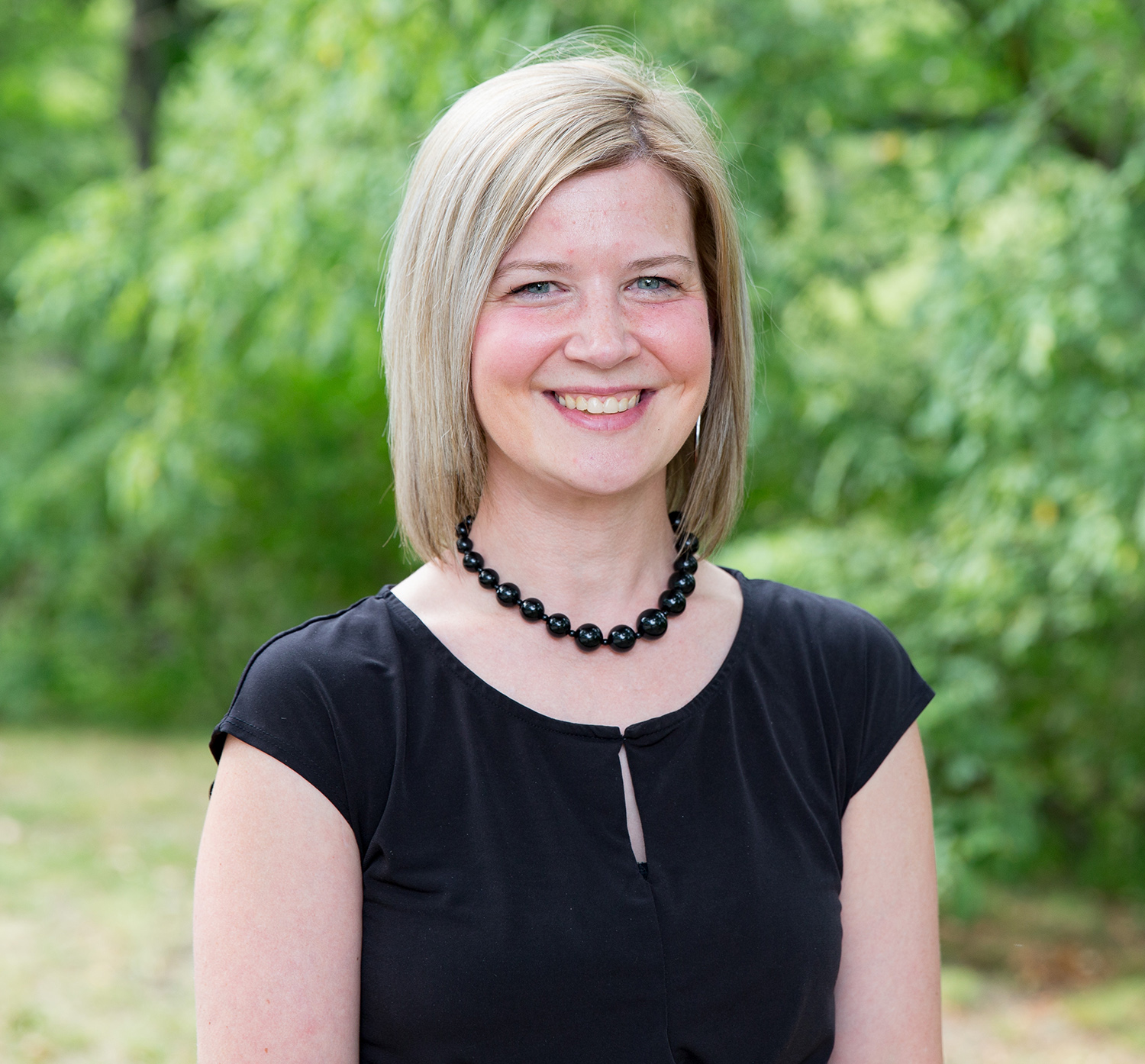 Lori Bruner smiles for the camera while standing in an outdoor setting. Bruner wears a short-sleeved black shirt and a black-beaded necklace. Bruner has shoulder-length blonde hair.