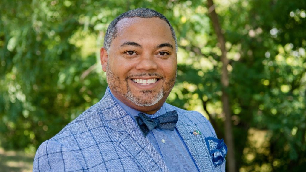 Bryan Beverly smiles for the camera in an outdoor setting. Beverly wears a blue button-down shirt, a navy blue bow tie and a plaid blue blazer. Beverly has black and gray hair cropped close to the head and subtle facial hair.