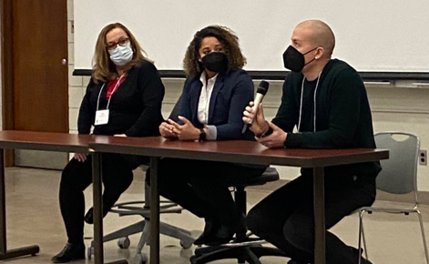 Three scholars sit side-by-side at a table. One holds a microphone to their mouth while speaking. All three wear face masks in various colors and patterns. 