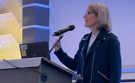 Nicole LaVoi speaks into a microphone at the Midwest Sport and Exercise Psychology Symposium conference. LaVoi wears a black blazer and a white top. LaVoi has glasses. LaVoi has shoulder-length blonde hair.
