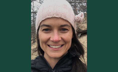 Rebecca Sandee headshot anchored by dark green rectangles on either side. Sandee smiles for the camera in an outdoor setting while wearing a dark top and a pink winter hat.