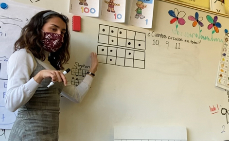 Mak Perez stands near a white board and points to a problem drawn on the board in a black marker. Perez wears a white shirt under a gray dress. Perez has shoulder-length, wavy brown hair.
