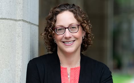 Tanya Wright smiles while leaning against a concrete building pillar. Wright wears a coral colored blouse under a black cardigan and dark-rimmed glasses. Wright has shoulder length curly brown hair.