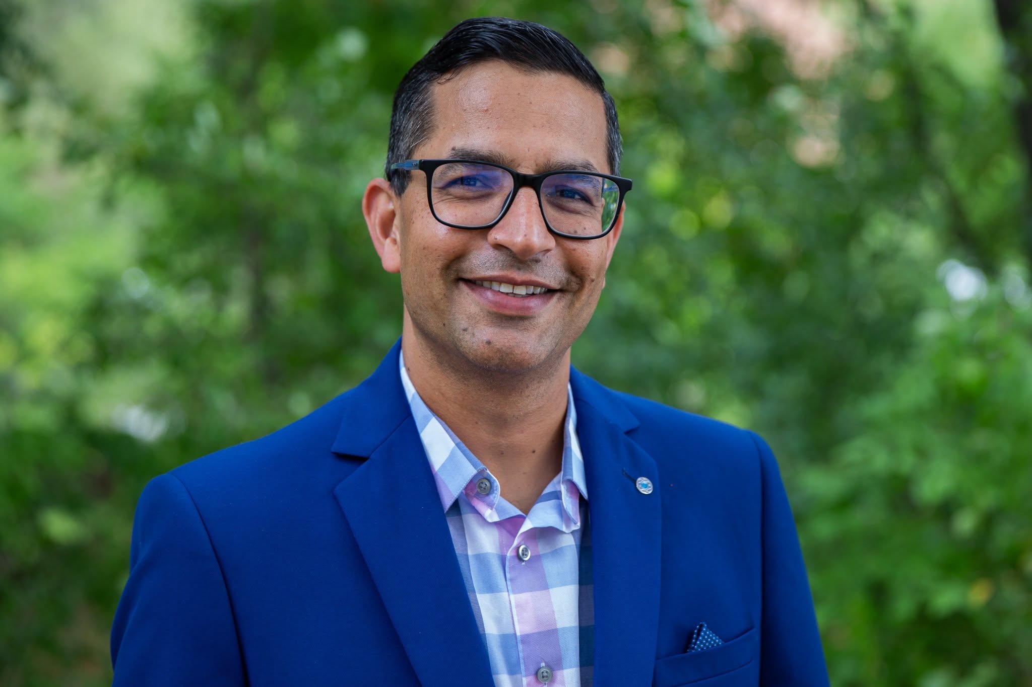 Aman Yadav smiles while standing outside in front of lush greenery. Yadav wears a blue and pink plaid shirt under a bright blue blazer. Yadav also wears dark-rimmed glasses. Yadav's black hair is short.