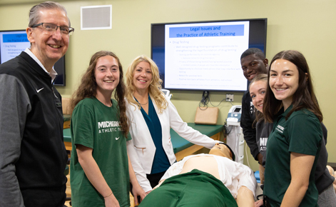 Spartan instructors and athletic training students smile for the camera in an athletic training classroom, surrounding a teaching mannequin used for teaching purposes.