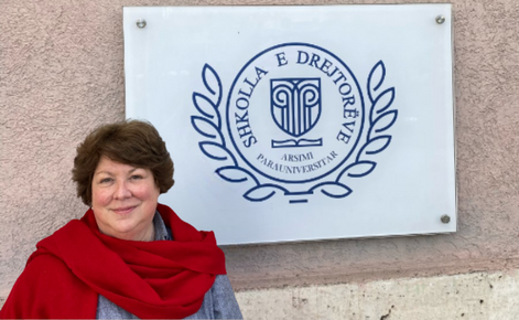 Smith smiles while standing in front of a building sign. Smith wears a gray top and a large red scarf. Smith has short brown hair. The sign behind Smith is primarily white and has navy blue lettering and logo.