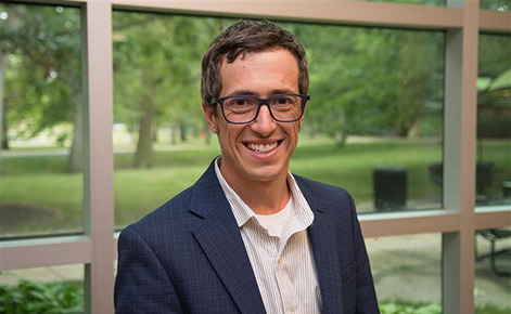 Matt Brodhead poses for a photo while standing in front of horizontal, rectangular windows in Erickson's Hall. A grassy courtyard is visible in the background. Brodhead wears a button down with pastel stripes under a dark blue blazer and dark-rimmed glasses. Brodhead has short, brown hair.