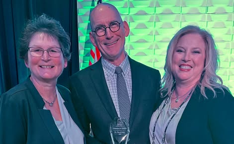 Three individuals pose for a photo at a conference, including David Arsen, MSU professor emeritus, at the center wearing a suit and a jacket and glasses.