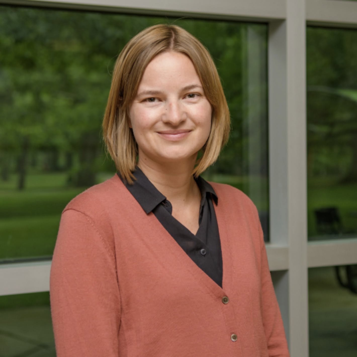 Alexandra Lee smiles for the camera against a row of horizontal, glass windows in Erickson Hall. Lee wears a navy blue button-down shirt under a coral blouse. Lee's blonde hair is about chin-length.