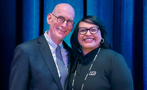 Two individuals smile toward the camera in a conference-like setting. The man wears a white button-down, gray tie and black blazer. The man also wears glasses. The woman wears a black top and a lanyard string for the conference, black-rimmed glasses and silver earrings. The woman has shoulder-length black hair.