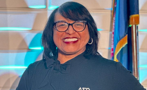 Monica Merritt smiles toward the camera while standing in a conference-room setting. Monica wears a black top, dark-rimmed glasses and silver hooped earrings. Monica has shoulder-length black hair.