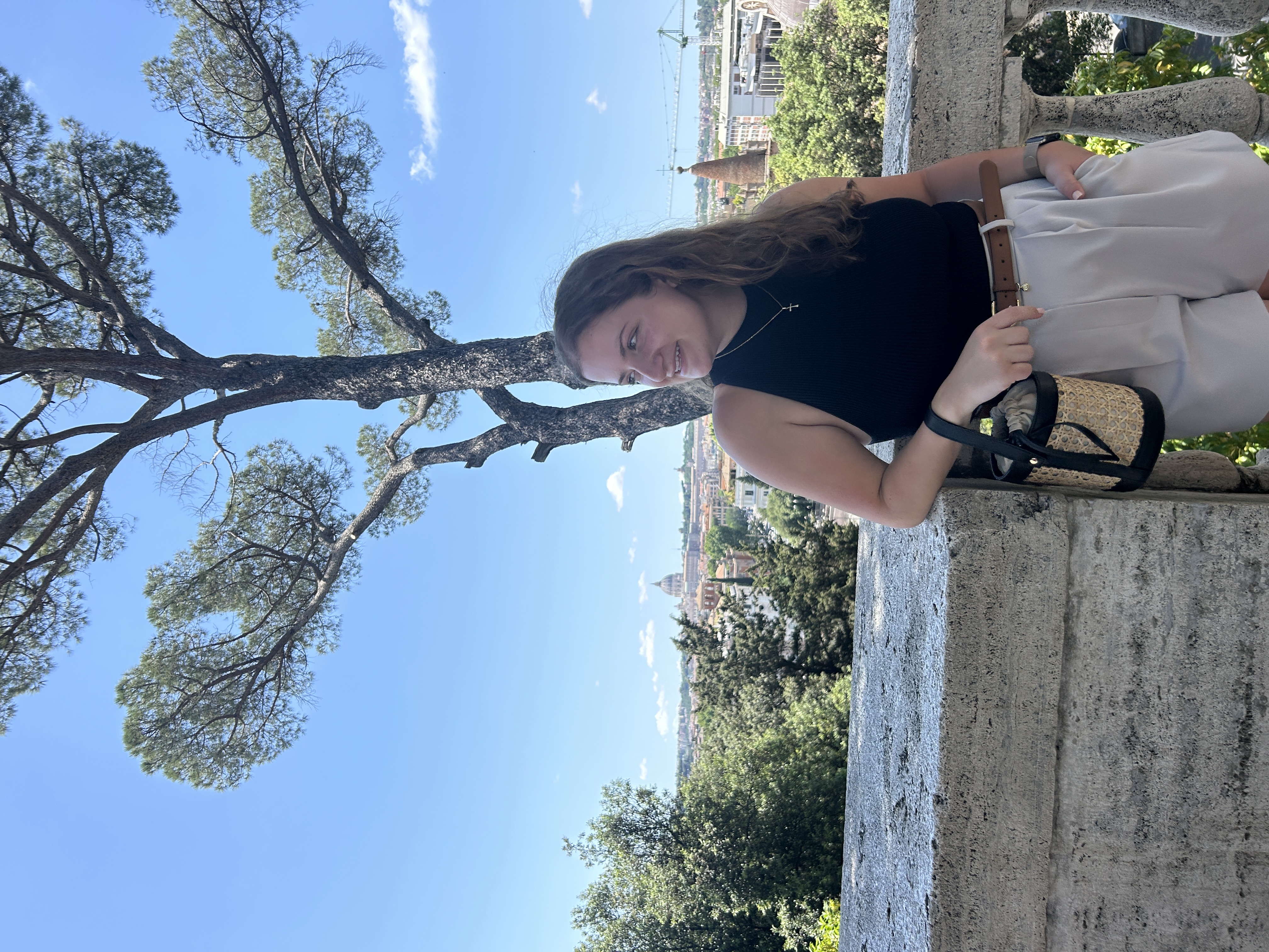 Sophia Amori stands outside and looks off camera. Amori is in an areas with lots of deciduous trees, and is standing in front of a stone ledge.
