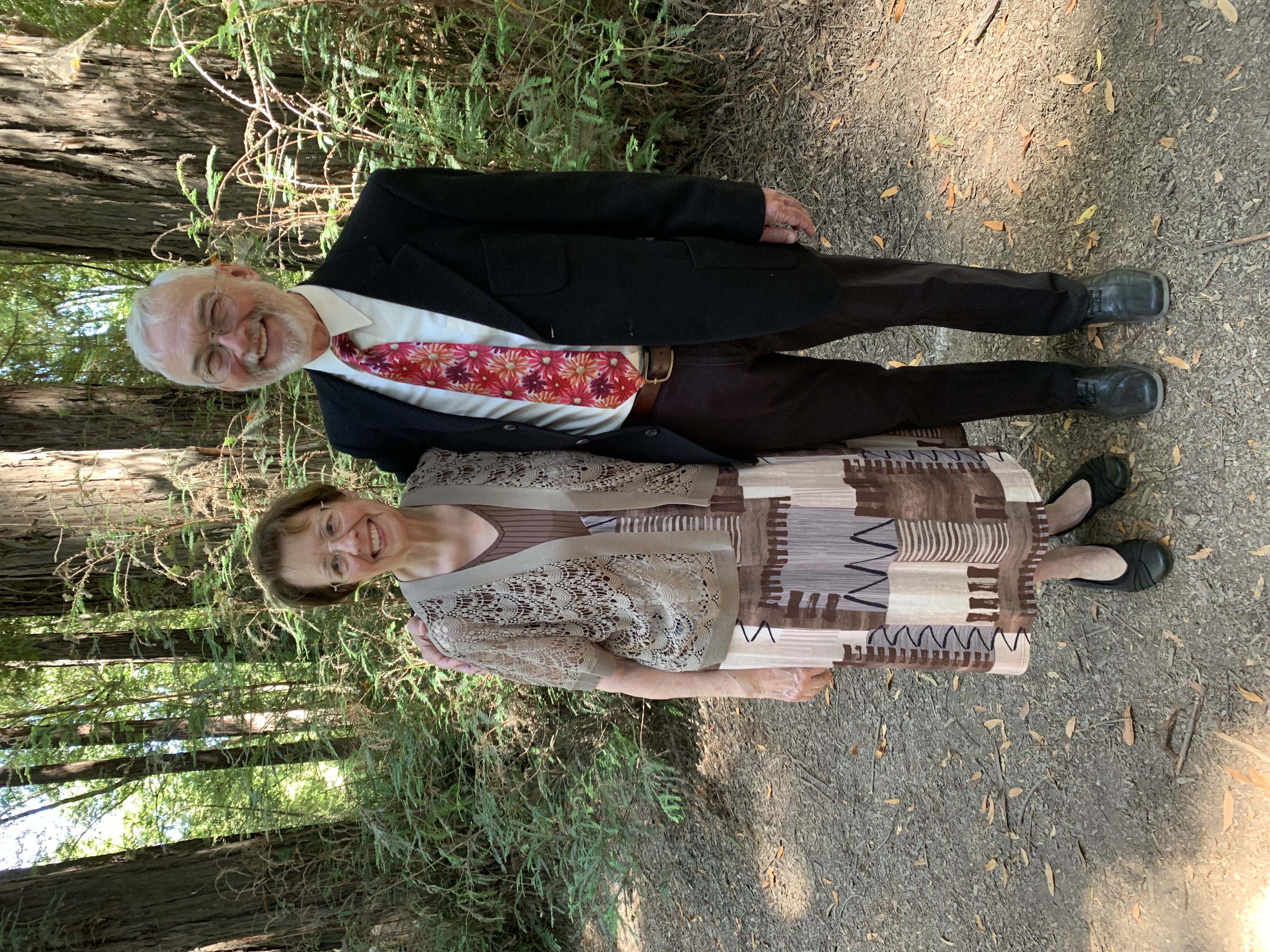 Two individuals stand side-by-side on a dirt path in a forest setting. One wears a brown dress with varying patterns and brown shoes; the individual has short brown hair and glasses. The other wears a dark suit and red tie and has white hair.