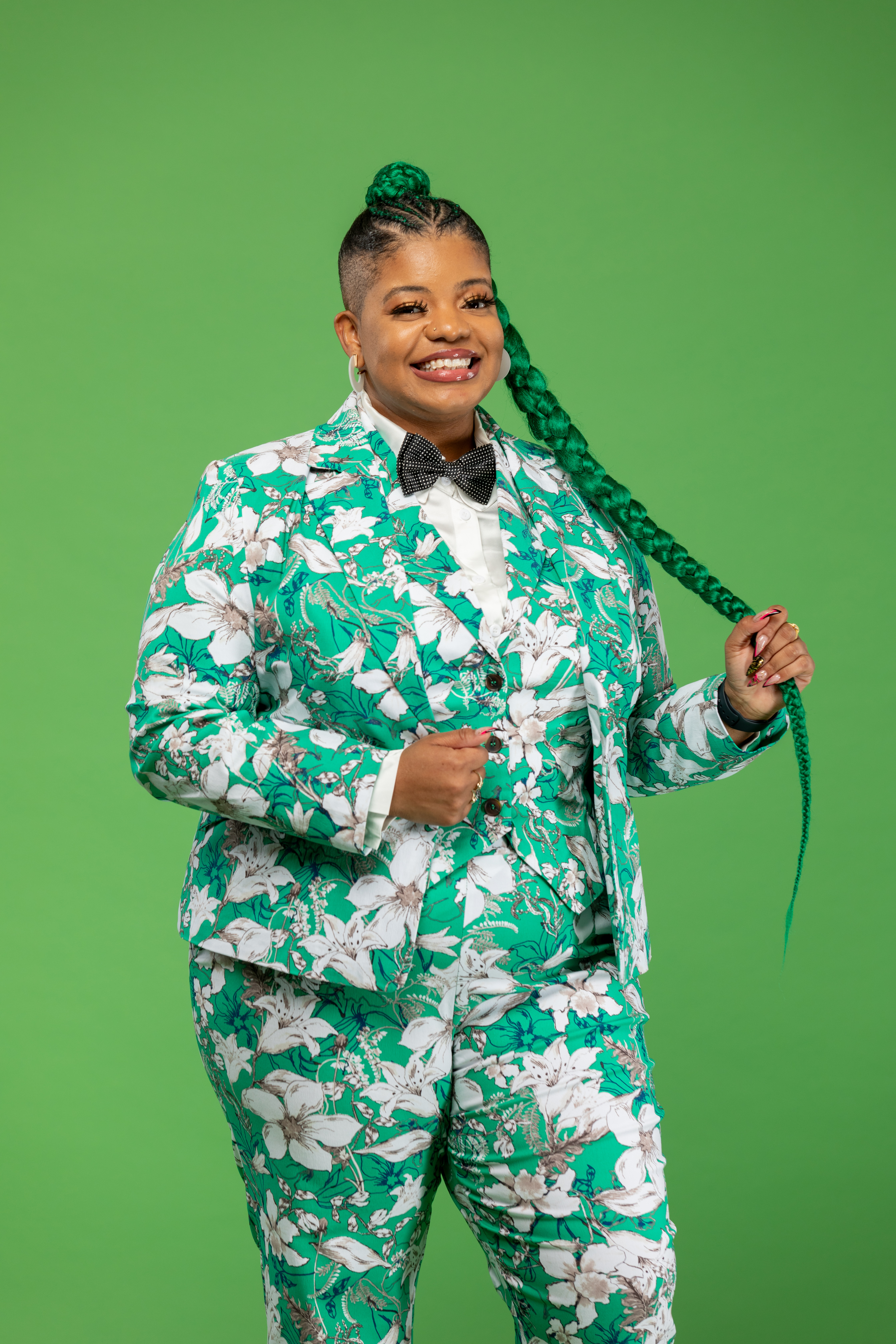 Kirby Gibson headshot; Kirby poses for the camera while standing against a green background. Kirby wears a green and white floral suit and a black bow tie. Kirby's black hair is long and in a braid.