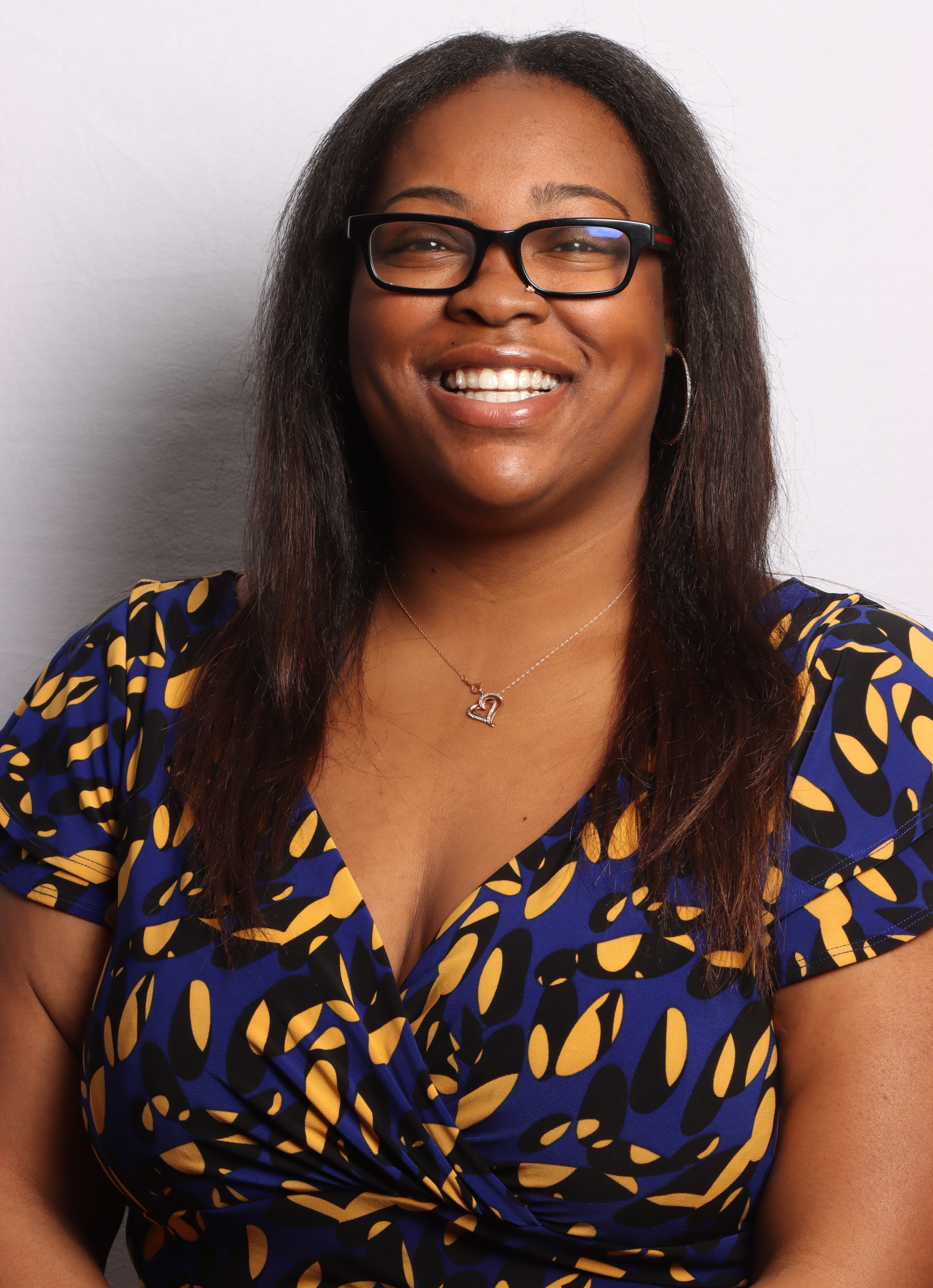 Tiyanna Peterson headshot. Tiyanna poses for the camera against a neutral white background. Tiyanna wears a blue, yellow and black patterned short-sleeve blouse and dark-rimmed glasses. Tiyanna's black hair is long and falls past the shoulders.
