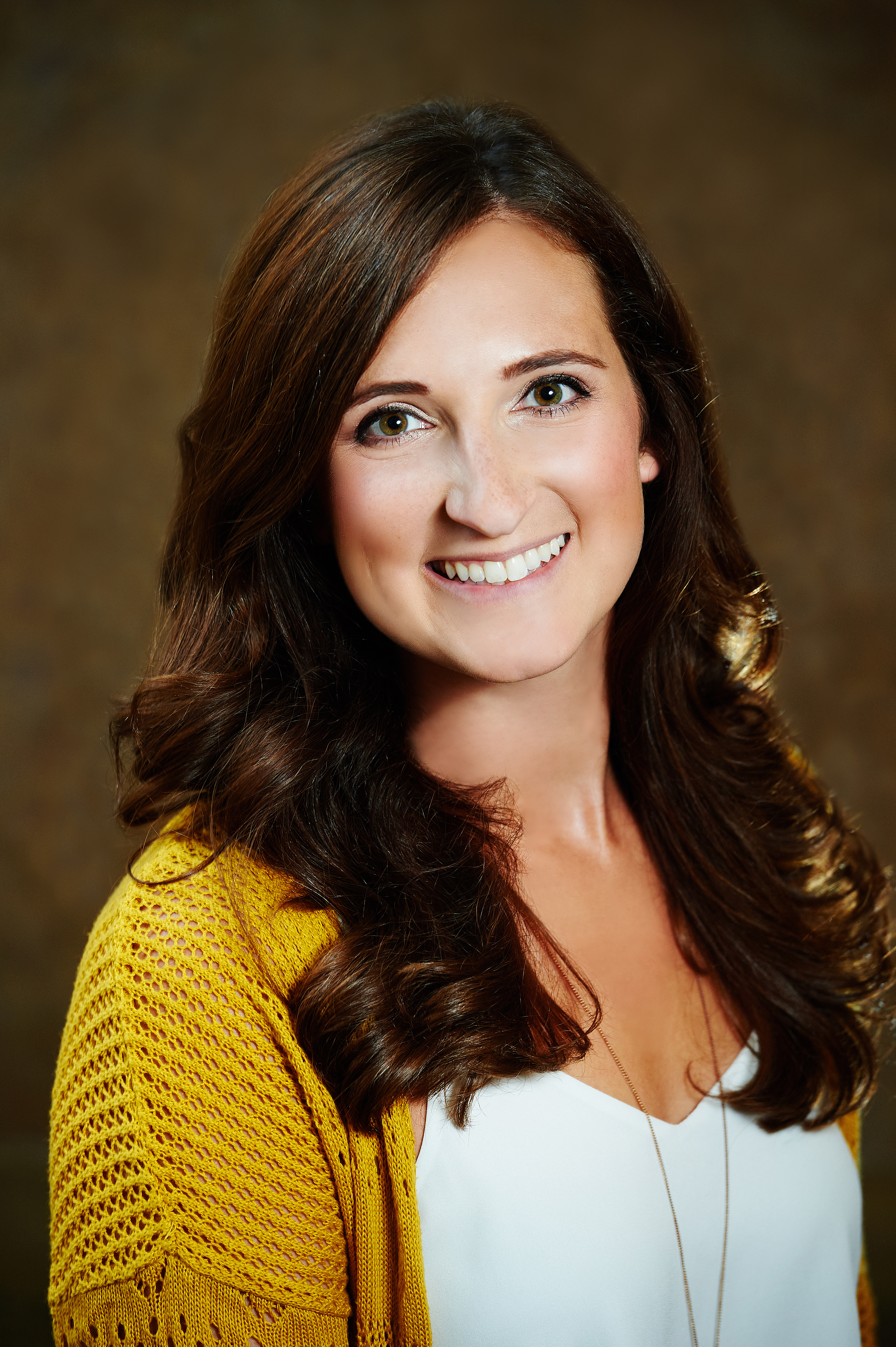 Sarah DiMaria smiles for the camera against a neutral brown background. DiMaria wears a white blouse under a yellow cardigan. DiMaria has long brown curly hair.